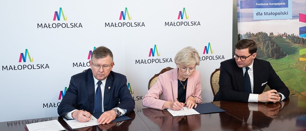 Kobieta i dwóch mężczyzn siedzą przy stole konferencyjnym. Jeden z mężczyzn i kobieta podpisują umowę. Wszyscy elegancko ubrani. Za nimi duża, biała plansza z napisami Małopolska. Obok planszy winder, plansza z napisem Fundusze Europejskie dla Małopolski.