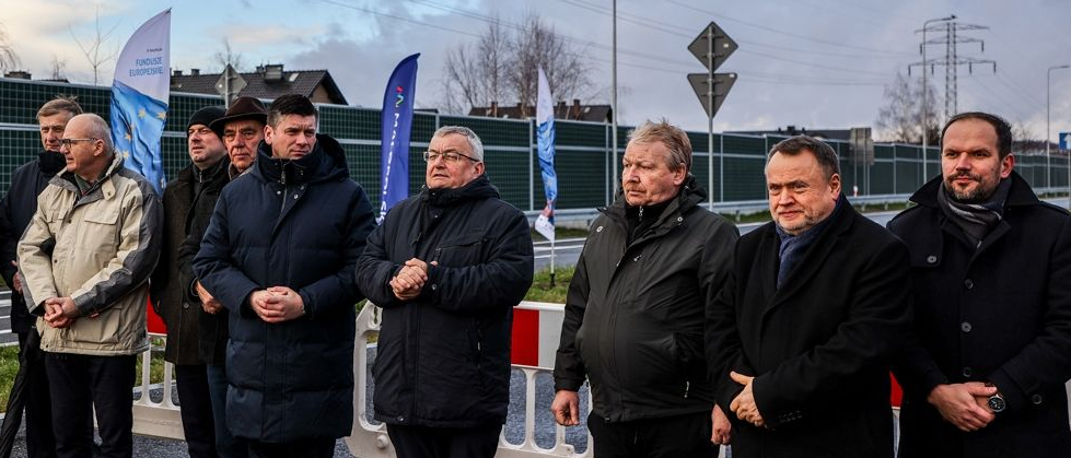 Kilku mężczyzn ubranych w zimowe kurtkii płaszcze stoi wzdłuż drogi. Za nimi widoczny fragment barierki drogowej oraz windery z logo Małopolski i Funduszy Europejskich. 