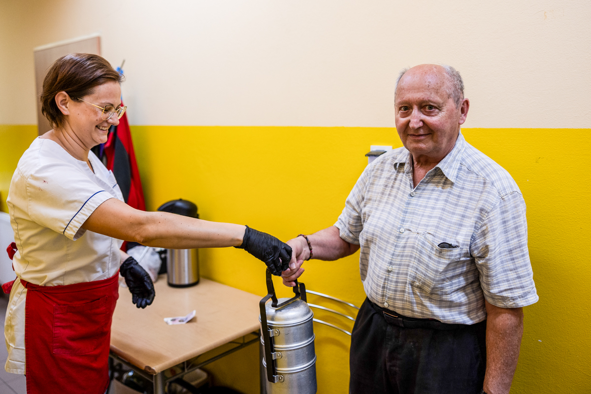 Pani w kuchni wręcza pojemnik z posiłkiem seniorowi.