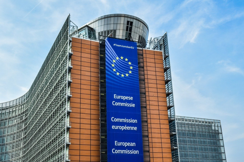 Budynek Komisji Europejskiej. Architektura budynku współczesna. Na środku zdjęcia pomarańczowa ściana z flagą UE i napisem Komisja Europejska. Po bokach, fasada ze szkła i stali w kształcie łuku.