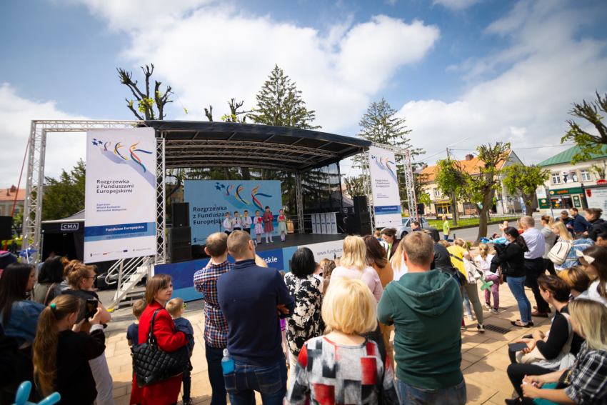 Na pierwszym planie tłum ludzi.. Na drugim planie zadaszona scena. Na scenie stoją dzieci, za nimi ustawiony jest duży ekran projekcyjny z wyświetloną grafiką.
