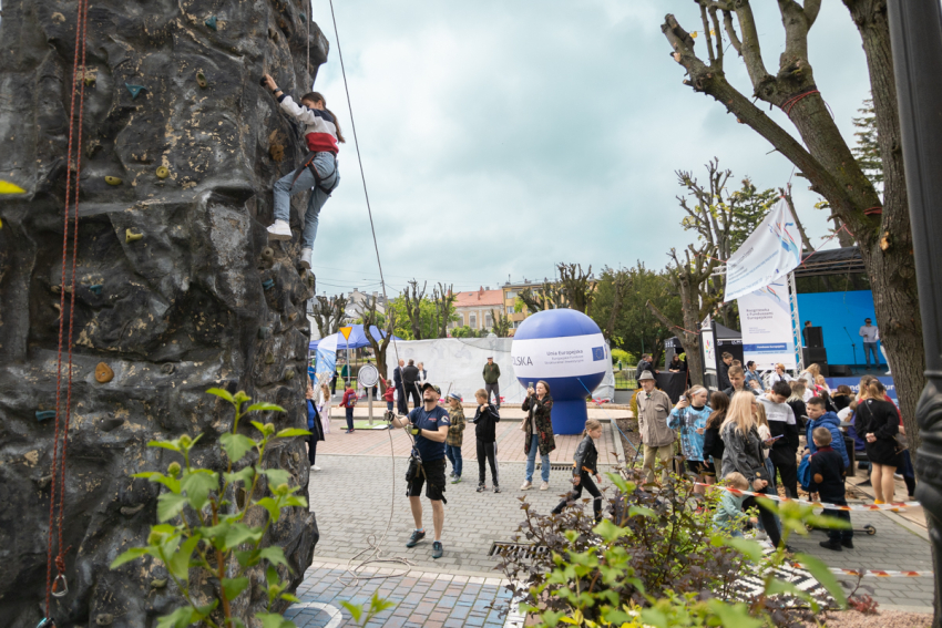 Po lewej stronie ścianka wspinaczkowa, po której wspina się młody człowiek. Na dole mężczyzna trzymający liny. Po prawej grupa ludzi oddzielona taśmą. W głębi scena i balon reklamowy.