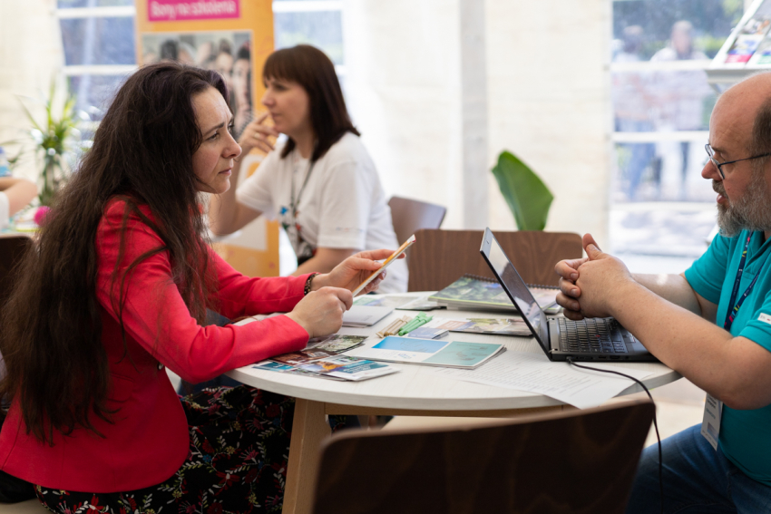 Kobieta i mężczyzna siedzą przy okrągłym stoliku, na którym znajdują się długopisy, ulotki i notesy oraz otwarty laptop. Kobieta trzyma w ręce ołówek.