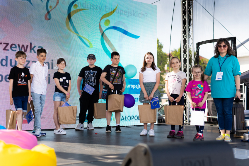 Dzieci z torbami prezentowymi ustawione w szeregu. Obok nich stoi kobieta i obejmuje jedno z dzieci. Za nimi znajduje się ekran projekcyjny z wyświetloną grafiką.