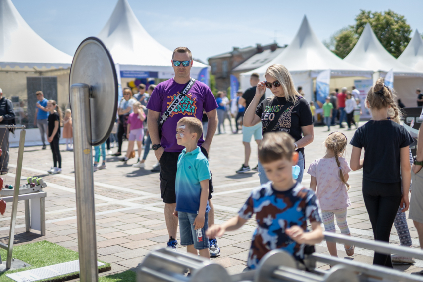 Metalowe urządzenia, przy których stoją dzieci i dorośli. Za nimi tłum ludzi i namioty o spiczastym zadaszeniu.