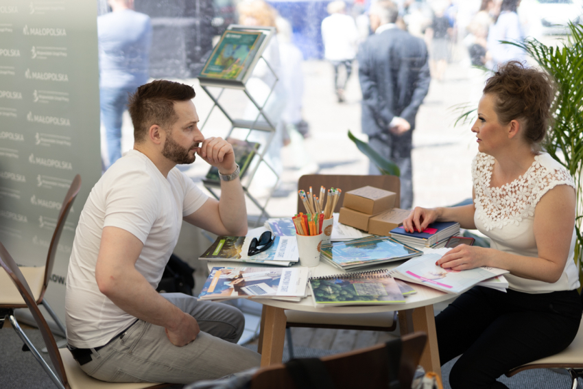 Mężczyzna i kobieta siedzą przy okrągłym stoliku, na którym znajdują się ulotki, biuletyny i notesy, a także kredki, ołówki i długopisy.