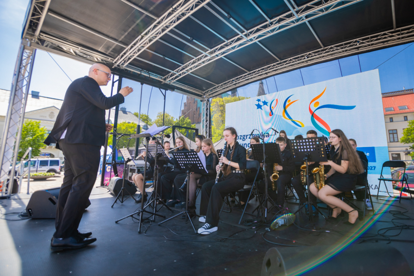 Zadaszona scena. Na pierwszym planie mężczyzna zwrócony w stronę grupy młodych ludzi, siedzących przy pulpitach z instrumentami w rękach. Na drugim planie duży ekran projekcyjny z wyświetloną grafiką.