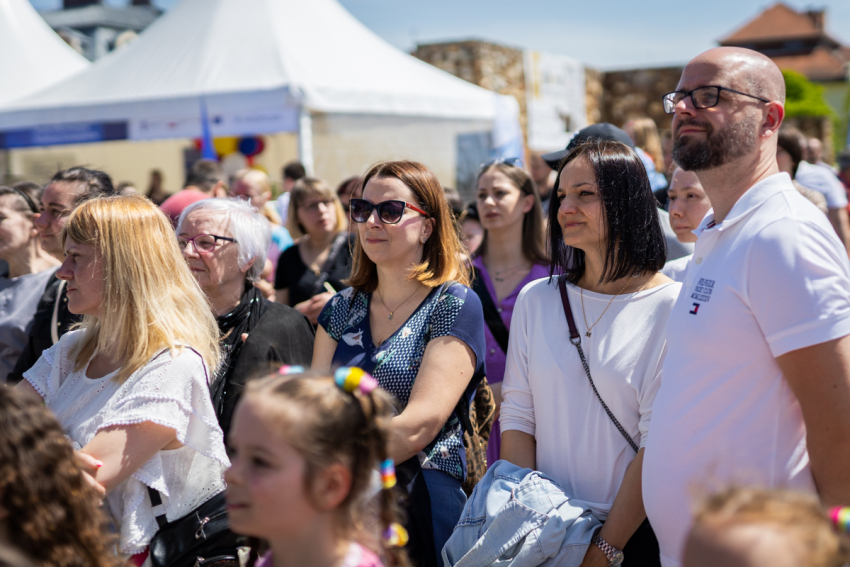 Grupa ludzi stoi i spogląda w jedną stronę. Świeci słońce. W tle namioty.