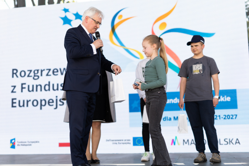 Elegancko ubrany mężczyzna trzymający w jednej ręce mikrofon, w drugiej torebkę prezentową. Obok niego stoi trójka dzieci. W tle duży ekran projekcyjny z wyświetloną grafiką dotyczącą Funduszy Europejskich.