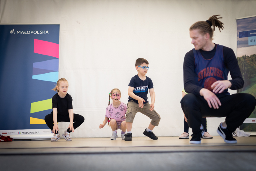 Mężczyzna z dredami kuca i spogląda na kucające obok dzieci. Za nimi dwa roll-upy, w tym jeden z napisem „Małopolska”