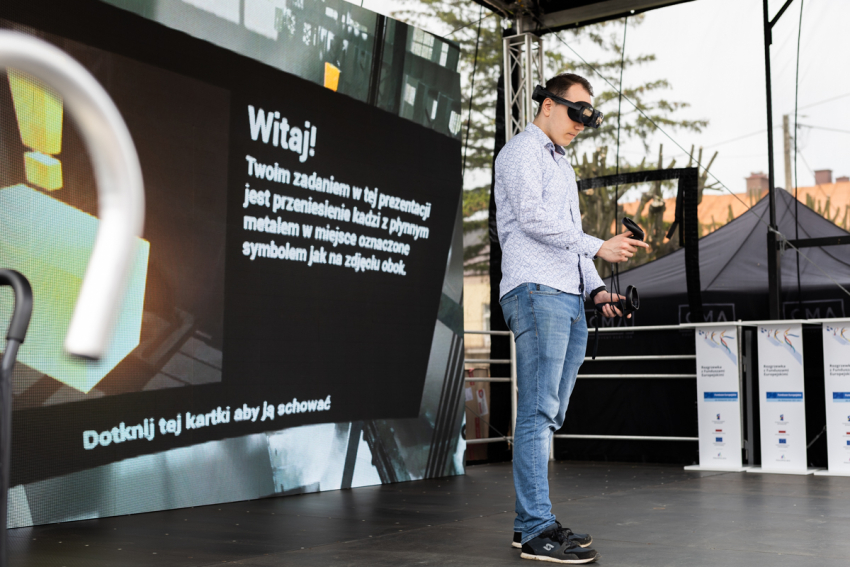 Mężczyzna w goglach do wirtualnej rzeczywistości i kontrolerami w obu rękach. Po prawej ekran projekcyjny z wyświetloną prezentacją.