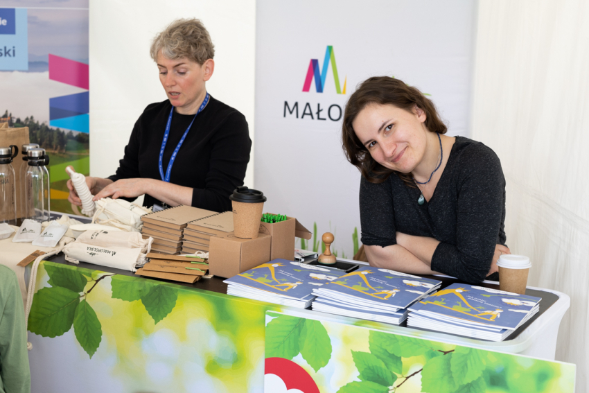 Dwie kobiety siedzą przy stole, na którym rozłożone są materiały promocyjne – biuletyny, notesy, długopisy, szklane bidony i kubek termiczny. Z nimi znajdują się dwa roll-upy – jeden z nich z logo Małopolski.
