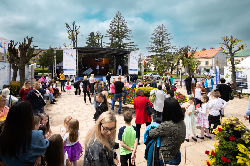 Tłum ludzi przed zadaszoną sceną. W tle kamienice, drzewa i pochmurne niebo.