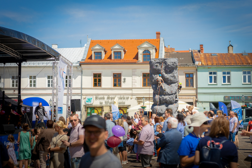 Na pierwszym planie tłum ludzi, scena z zadaszeniem oraz ścianka wspinaczkowa. Na drugim planie rząd piętrowych kamieniczek oraz bezchmurne niebo.