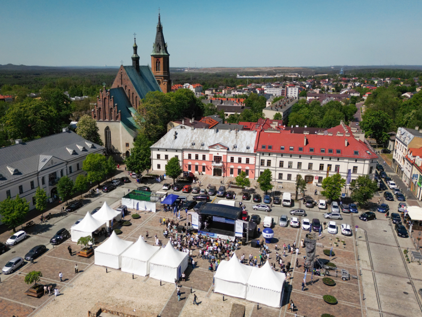 Widok z lotu ptaka na rynek miejski otoczony kamienicami. W centrum ustawione białe namioty imprezowe oraz scena z zadaszeniem. Przed sceną tłum ludzi, a za sceną zaparkowane samochody.