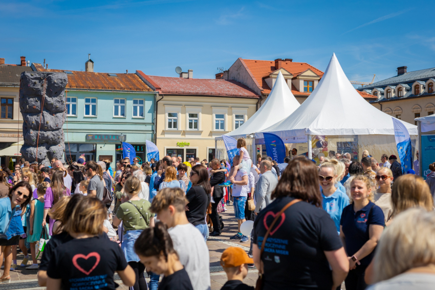 Na pierwszym planie tłum ludzi. Na drugim planie ścianka wspinaczkowa, namioty imprezowe ze spiczastym zadaszeniem oraz kilka flag reklamowych. W tle rząd kamienic i bezchmurne niebo.