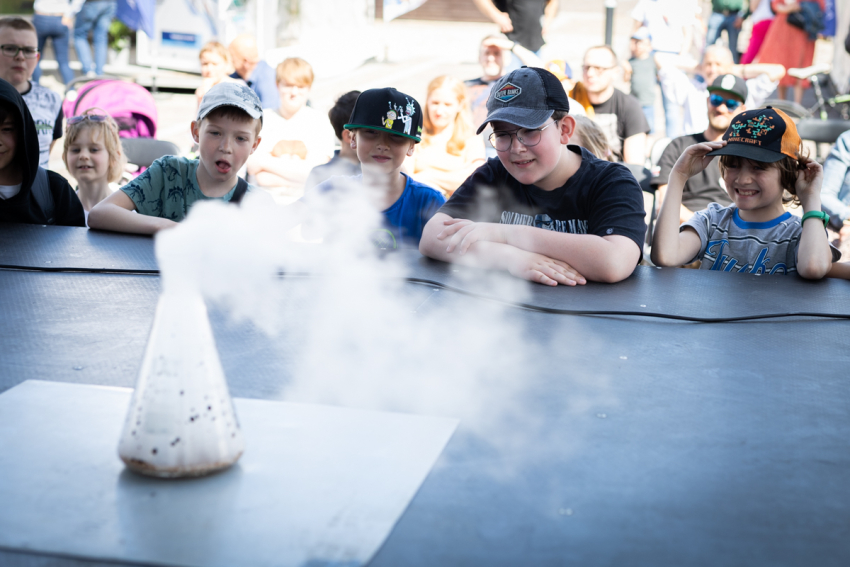 Grupka dzieci oparta o blat spogląda na szklane naczynie w kształcie stożka, z którego wydobywa się dym.