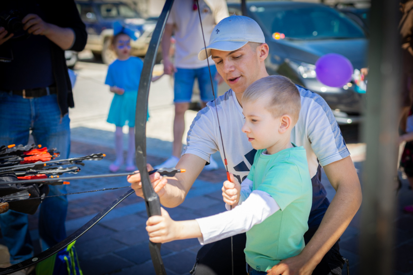 Kilkuletni chłopiec trzyma sportowy łuk. Obok kuca młody mężczyzna, który obejmując dziecko, podaje mu strzałę.
