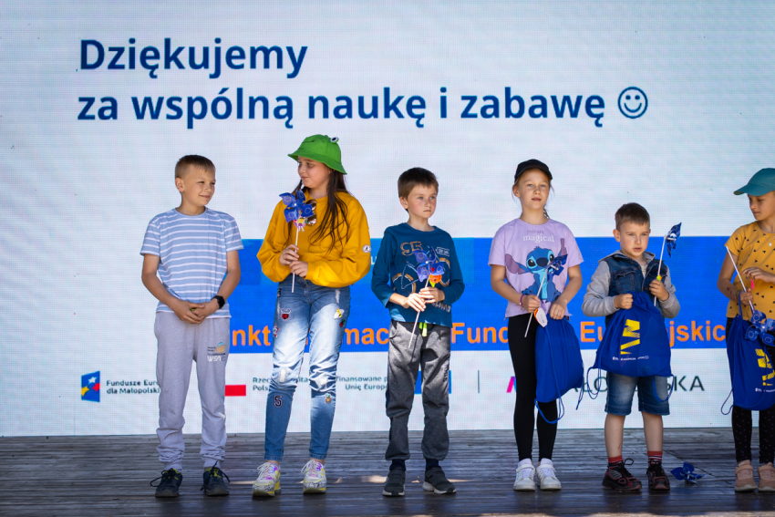 Grupka 6 dzieci w wieku wczesnoszkolnym stojących obok siebie na scenie. Niektóre z nich trzymają w rękach niebieskie wiatraczki z żółtymi gwiazdkami (Znak Unii Europejskiej). Za dziećmi w górnej części zdjęcia napis &quot;Dziękujemy za wspólną naukę i zabawę&quot;. Na dole widoczne fragmenty informacji o Funduszach Europejskich.