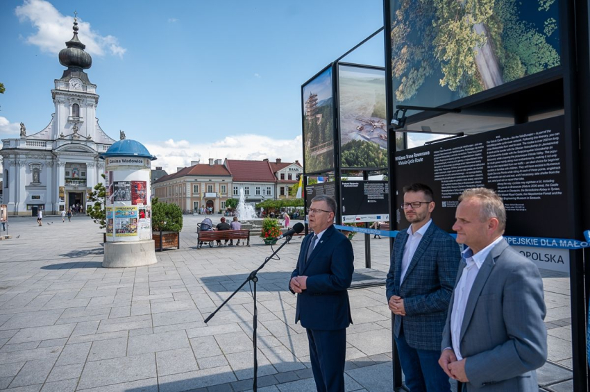Trzech mężczyzn, ubrani w białe koszule i marynarki stoją przy metalowej konstrukcji , na której umieszczone są zdjęcia dużego formatu przedstawiające piękne widoki z Małopolski. Jeden z nich przemawia do mikrofonu. Na dalszej części planu mniejsze kamieniczki, kościół, ławki, na których siedzą ludzie , kwiaty w donicach, fragment drzewa. Słoneczny dzień.