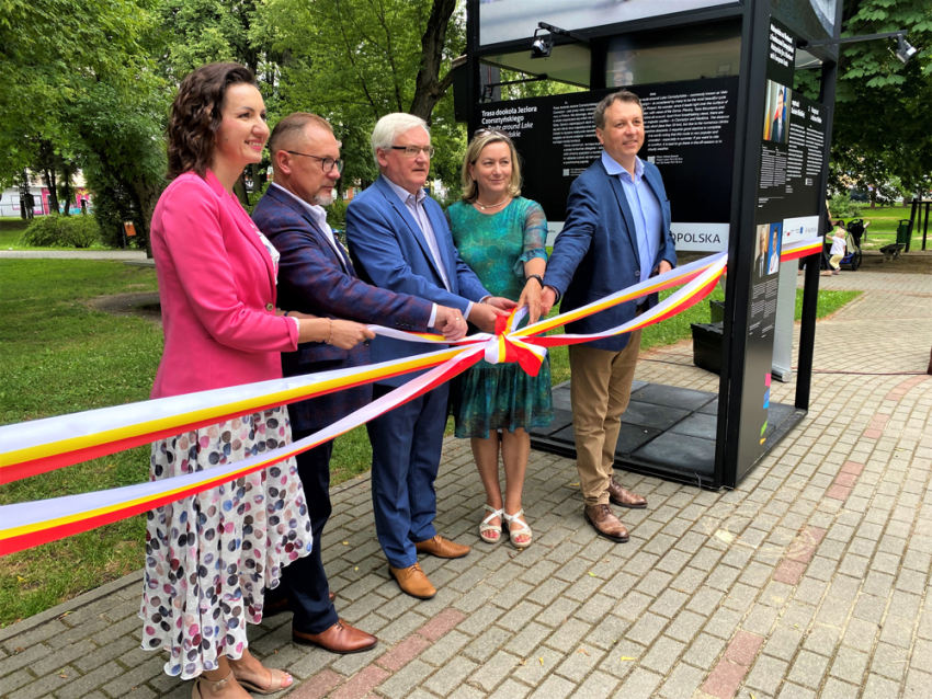 Dwie uśmiechnięte, elegancko ubrane kobiety i trzech mężczyzn w garniturach przecinają wstęgę na znak otwarcia wystawy o Małopolsce. Obok nich fragment konstrukcji, na której znajdują się zdjęcia inwestycji w Małopolsce powstałe dzięki Funduszom Europejskim. Za nimi widoczny park, trawnik, drzewa, ławka i bawiące się małe dziecko.