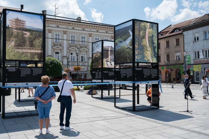 Starsza kobieta i starszy mężczyzna oglądają ogromne zdjęcia, które przedstawiają inwestycje zrealizowane przy wsparciu Funduszy Europejskich. Na pierwszym planie zdjęcie wieży widokowej w Krynicy i trasy rowerowej nad Wisłą. W tle kilka osób, stare ale i odnowione kamieniczki, trochę zieleni, korona drzewa, kwiatki. Ciepły, słoneczny dzień.