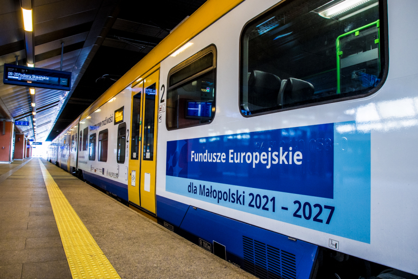 pociąg Nowoczesny pociąg - biało, żólto, niebieski stoi przy peronie , na stacji. Na widocznym wagonie napis: Fundusze Europejskie dla Małopolski 2021-2027. Na górze po lewej stronie wisi tablica z wyświetloną nazwą stacji. Peron jest oświetlony. Pora dnia noc.