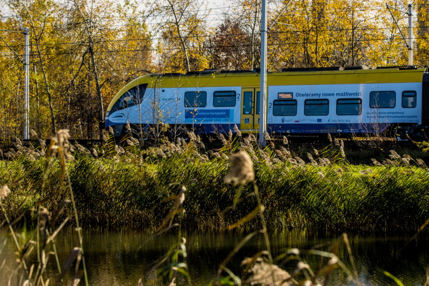 pociąg Żółto, biało, niebieski pociąg z napisami : Otwieramy nowe możliwości, Fundusze Europejskie. Pociąg przejeżdża przez las lub park -drzewa, trawy, widoczny fragment rzeki lub stawu. Słoneczny dzień.