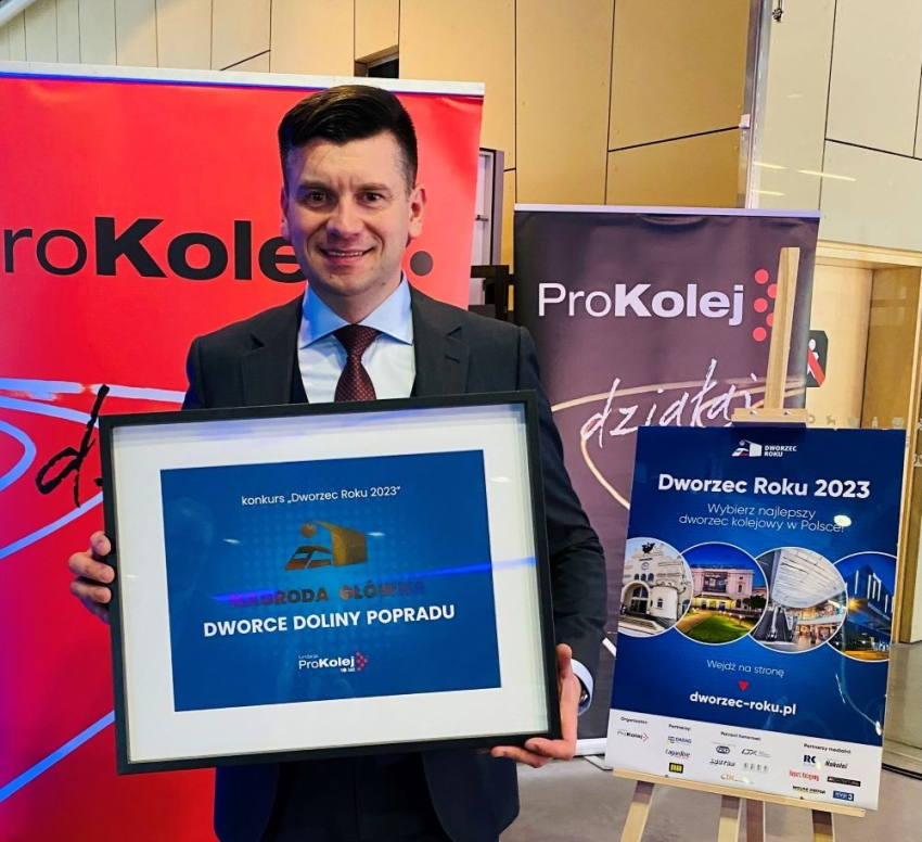 Uśmiechnięty mężczyzna w garniturze trzyma oprawioną w ramkę, pisemną nagrodę za Dworzec Roku. Za nim sztalugi z plakatami informującymi o konkursie oraz widoczne logo ProKolej. 