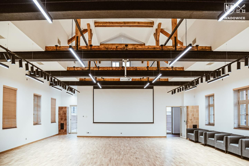 fot. Urząd Miejski w Wadowicach Poczekalnia dworca PKP. Jasna podłoga, białe ściany i sufit, widoczna drewniana konstrukcja z belek przy suficie. Po obu stronach sali okna z żaluzjami, po prawej rząd szarych puf, na wprost, po obu stronach drzwi.