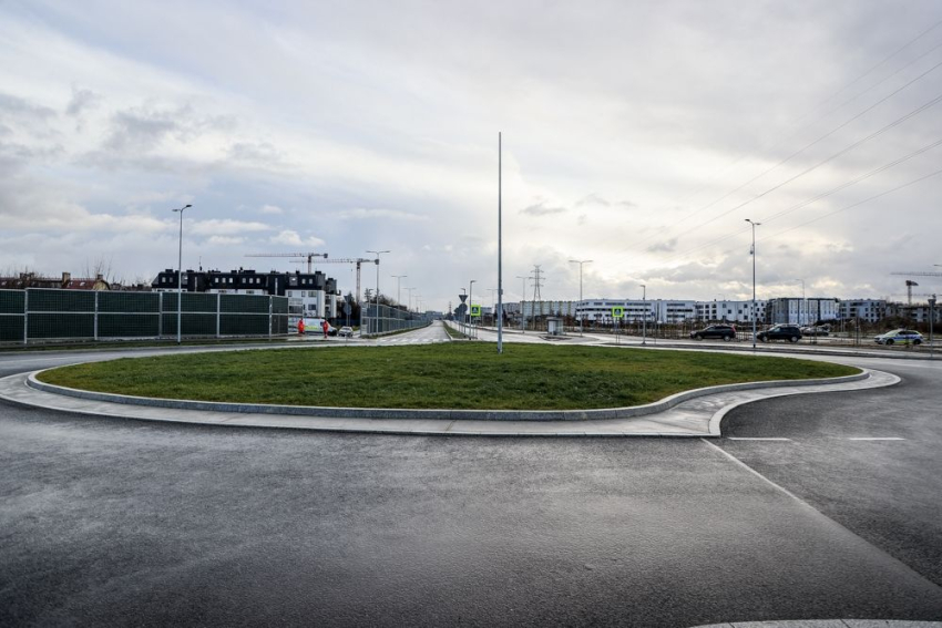 Rondo. W środku ronda zielona trawa. W oddali po obu stronach widoczne bloki. Część z nich w budowie, widoczne dźwigi, samochody osobowe.