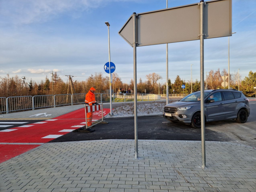 Fragment mostu. Po lewej stronie mężczyzna w pomarańczowym kombinezonie roboczym, stoi przy ścieżce rowerowej i przestawia płotek drogowy. Po prawej szary samochód osobowy. Widoczny drogowskaz od tyłu. W oddali widoczne drzewa.