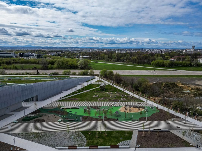 Nowoczesny budynek Centrum Nauki oraz przestrzeń wokół niego widziana z góry. Budynek główny, drogi do niego prowadzące, park wokół, oddalone inne budynki, zieleń, drzewa, pogodne niebo usłane chmurami.