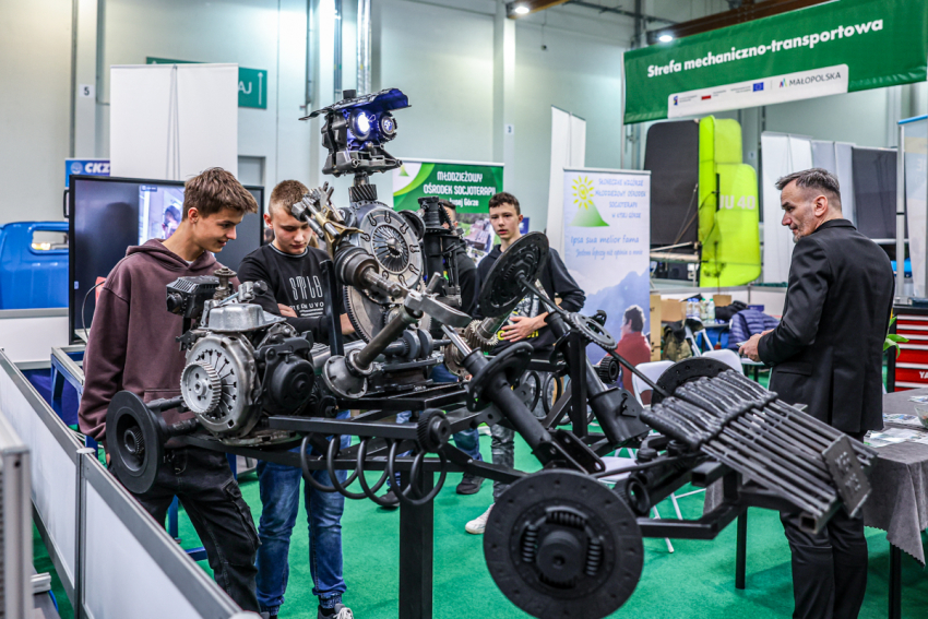 Stoisko mechaniczno-transportowe. Szkielet/konstrukcja samochody wyścigowego. Wokół kilka młodych osób oraz starszy pan.