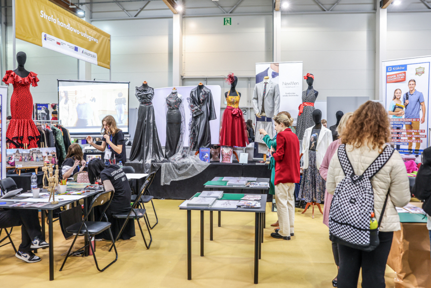 Stoisko krawieckie. Stoliki, na których leżą magazyny o modzie i projekty ubrań. Scena, na której stoją manekiny ubrane w eleganckie suknie. Przy stolikach kilka osób.