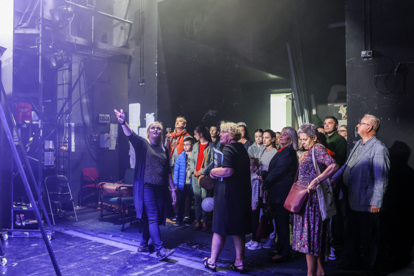 Kulisy teatru. Kobieta pokazuje grupie osób sprzęty się tam znajdujące.
