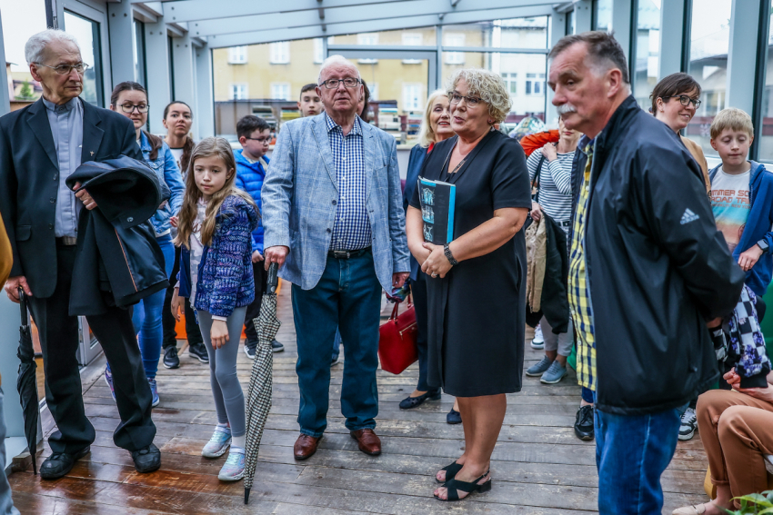 Grupa osób, w różnym wieku stoi w przeszklonym pomieszczeniu.