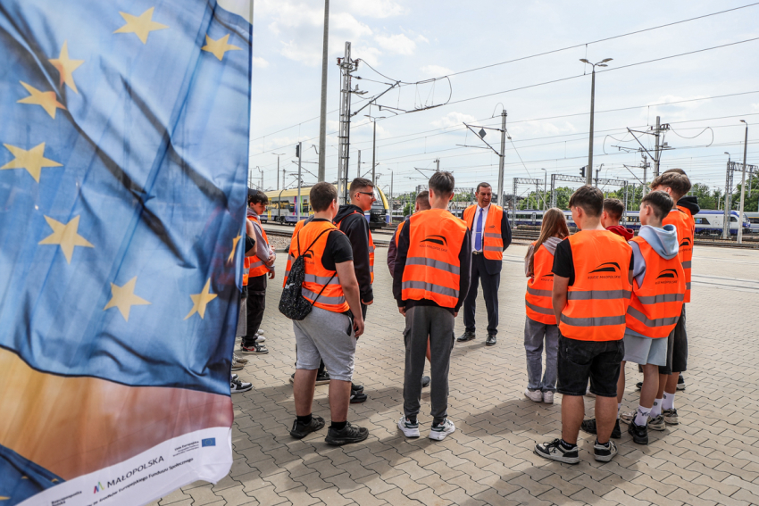 Grupa dzieci szkolnych i mężczyzna, w kamizelkach odblaskowych na peronie kolejowym.