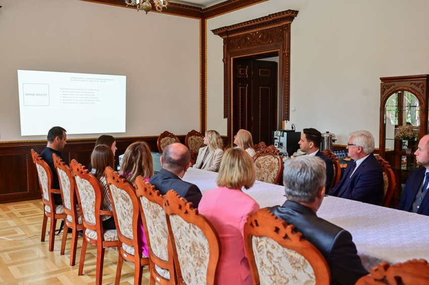 Grupa osób siedzi przy stole konferencyjnym i oglądają prezentację na ekranie rzutnika.
