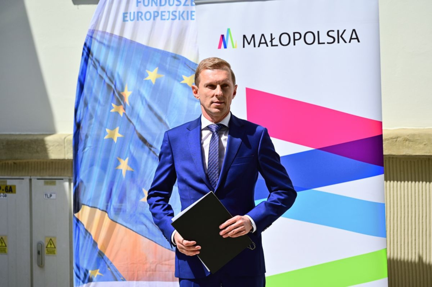 Młody mężczyzna stojący na zewnątrz. Za nim windery Fundusze Europejskie i Małopolska.