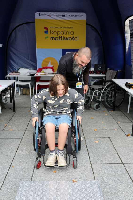 Dziecko na wózku inwalidzkim. Za nim instruktor, który tłumaczy działanie sprzętu.