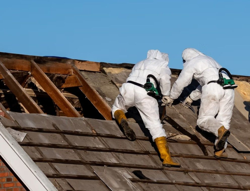 Robotnicy w kombinezonach ochronnych usuwają azbest z dachu.