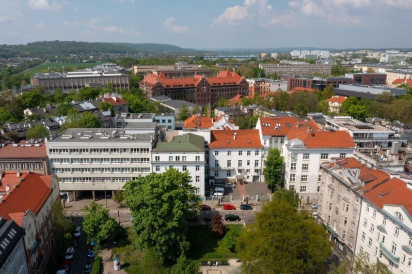 Budynek szpitalny w otoczeniu kamienic i zieleni.