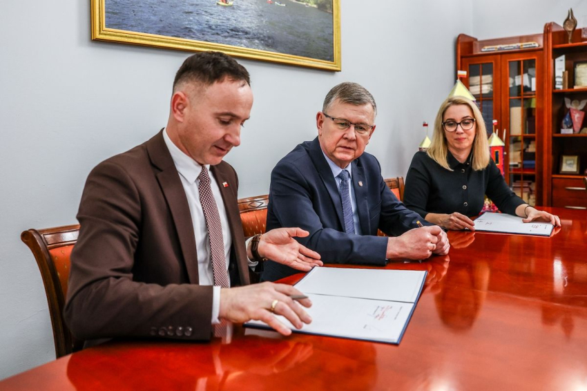 Mężczyzna podpisujący umowę. Za nim wicemarszałek Witold Kozłowski i kobieta w blond włosach.