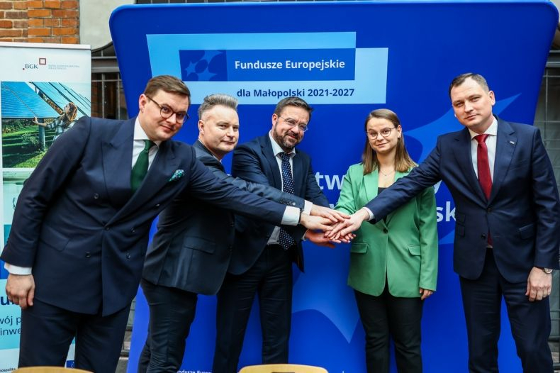 Pięć osób w formalnych strojach stoi przed niebieskim tłem z napisem „Fundusze Europejskie dla Małopolski 2021-2027”. Wyciągają ręce w centralnym stosie, pokazując gest współpracy.