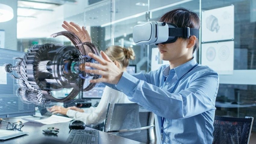 Mężczyzna w okularach VR pracuje z trójwymiarowym modelem holograficznym, wykonując gesty. W tle biuro inżynierskie oraz inni pracownicy.