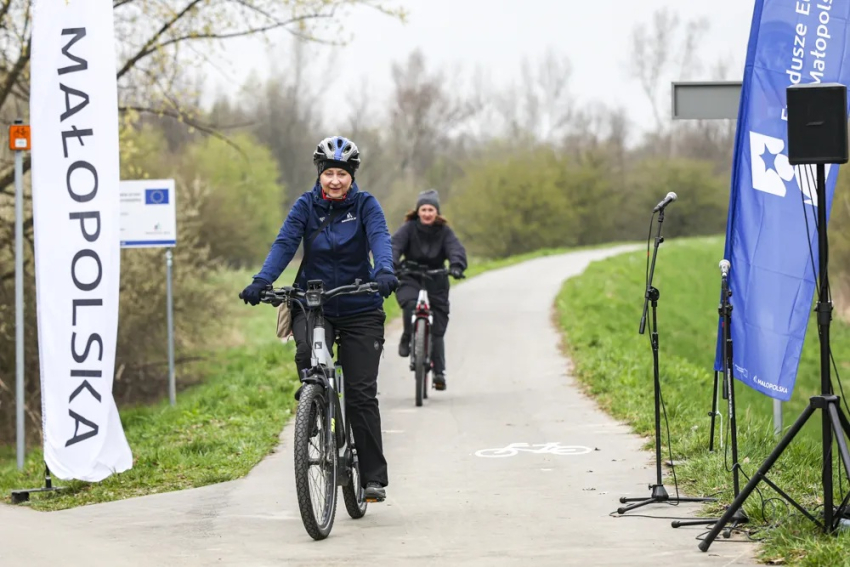 Rowerzyści jadą po asfaltowej ścieżce; po lewej widoczna flaga Małopolski, po prawej flaga Funduszy Europejskich