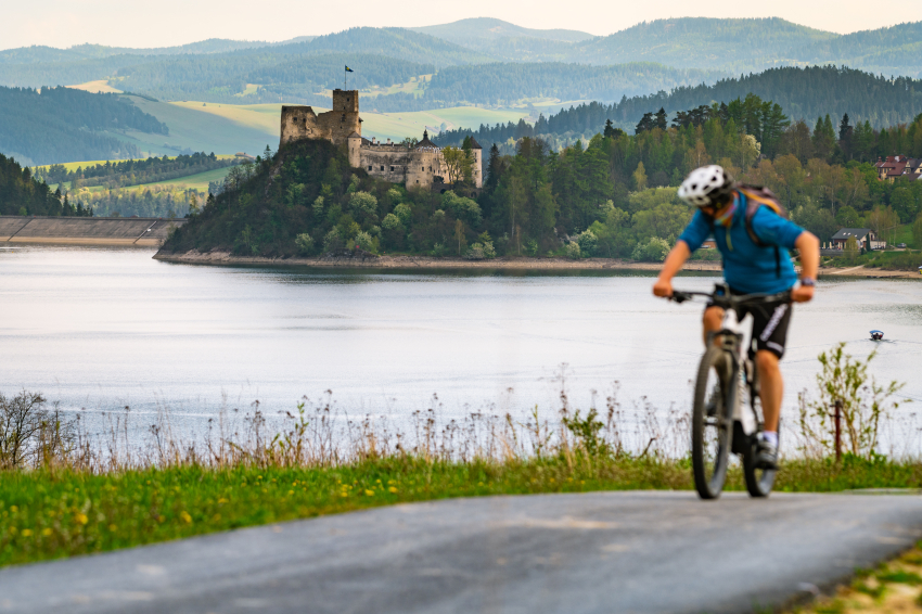 Mężczyzna jadący na rowerze. W tle jezioro, zamek i góry