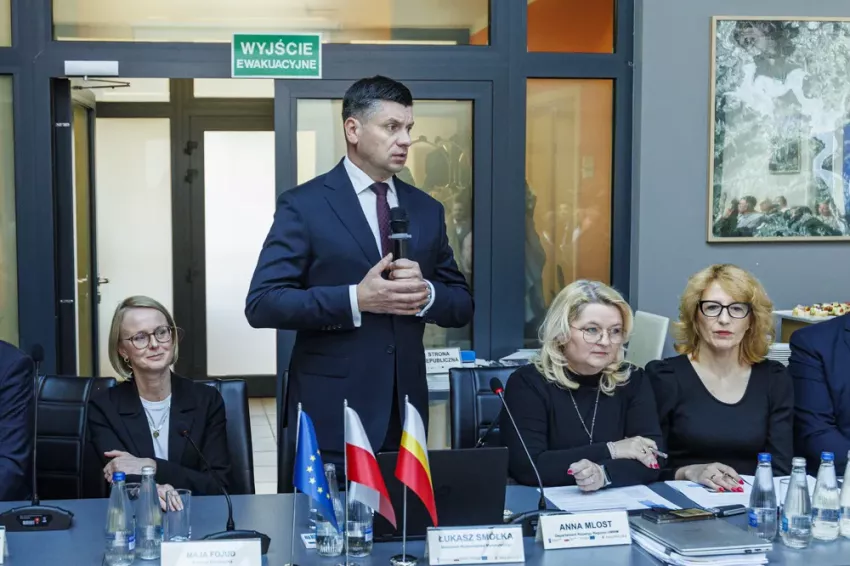 Marszałek Łukasz Smółka przemawia podczas posiedzenia Komitetu Monitorującego stojąc przy stole konferencyjnym, w otoczeniu uczestników wydarzenia