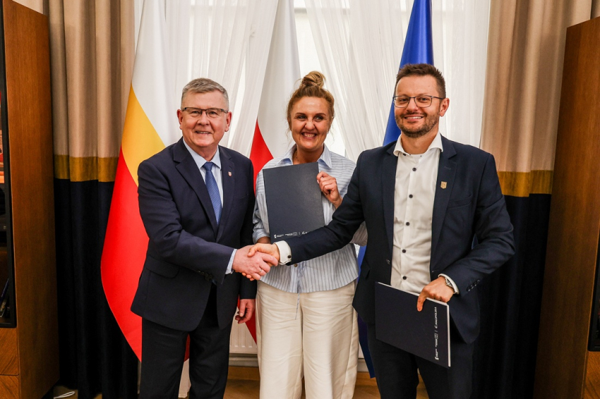 Witold Kozłowski i Bartosz Kaliński ściskają swoje dłownie tuż po podpisaniu umowy. Kinga Gurdek trzyma okolicznościową teczkę z dokumentami. Cała trójka stoi na tle flag Małopolski, Polski i Unii Europejskich
